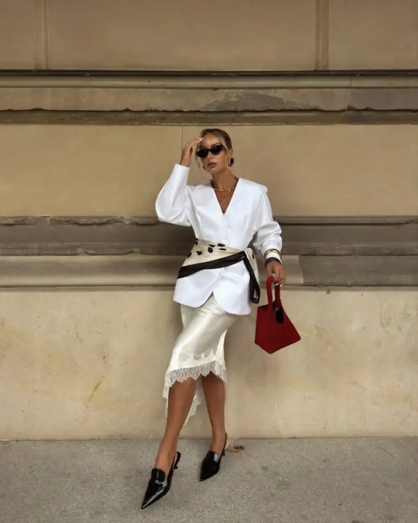 White blazer paired with an ivory satin skirt with lace trim, printed waist scarf, black slingback heels, and red handbag.