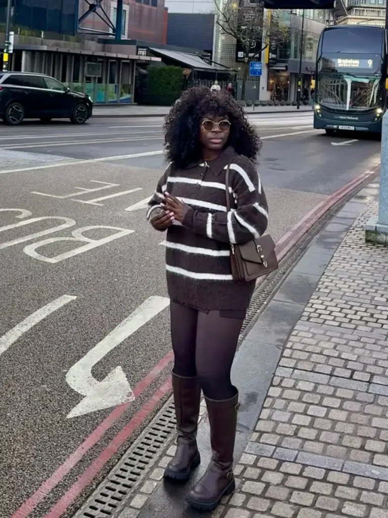 Black woman wearing an oversized chocolate brown and cream striped sweater dress with sheer tights, tall brown boots, sunglasses, and a structured brown shoulder bag for a cozy fall outfit.