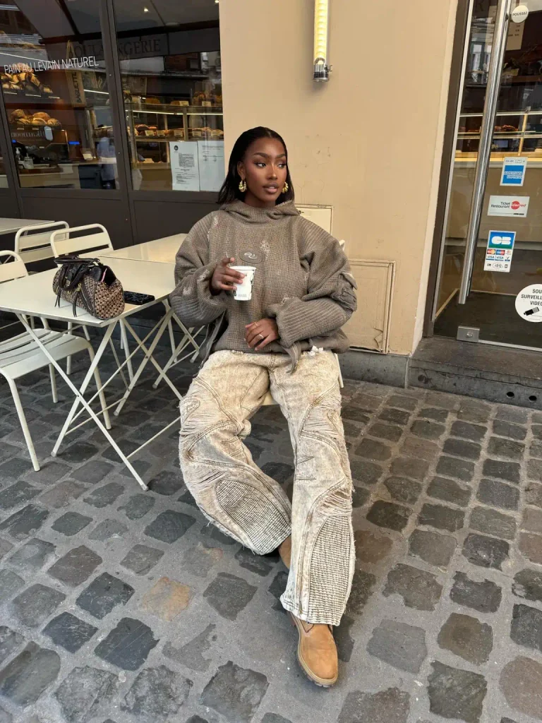 Black woman styling an oversized taupe distressed hoodie with baggy acid-wash cargo jeans, tan ankle boots, and gold statement earrings.