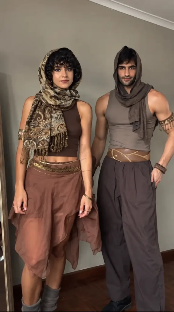 Couple wearing brown desert warrior costumes with wrapped scarves, loose layers, belts, and bronze accessories.