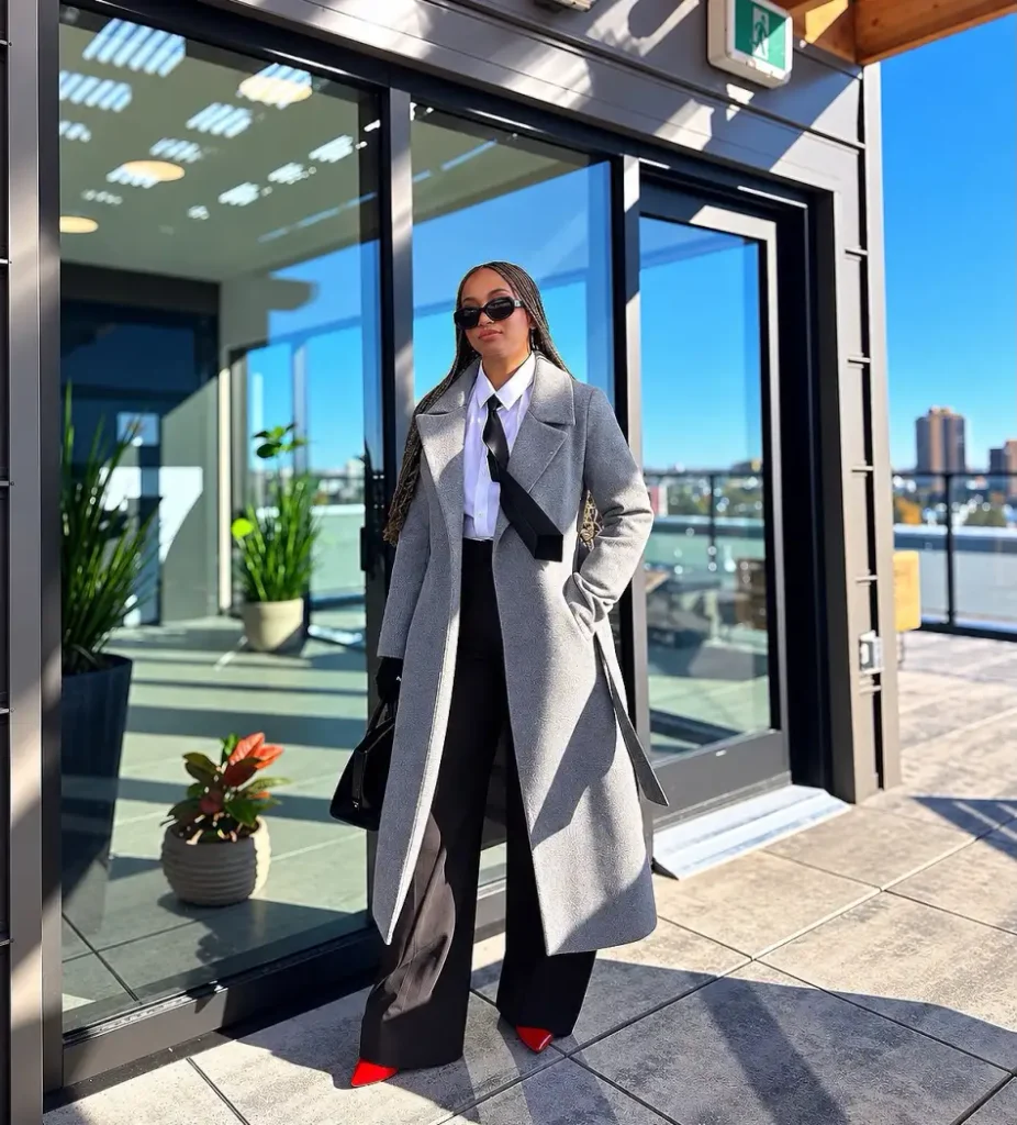Black woman wearing a long light gray coat over a white button-down shirt and black necktie with black trousers, red pointed heels, and dark sunglasses.