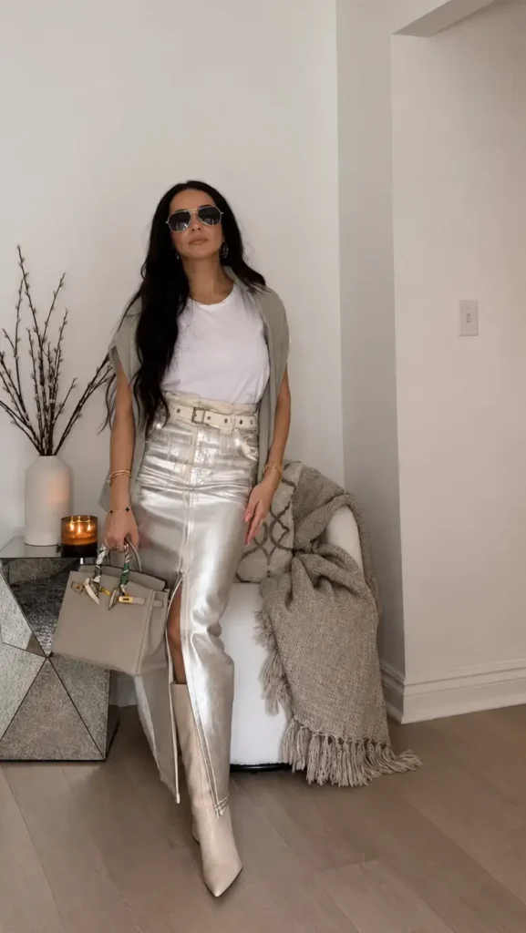 Petite woman wearing a white T-shirt with a metallic silver midi skirt, wide cream belt, neutral knit layer, and pointed cream boots.