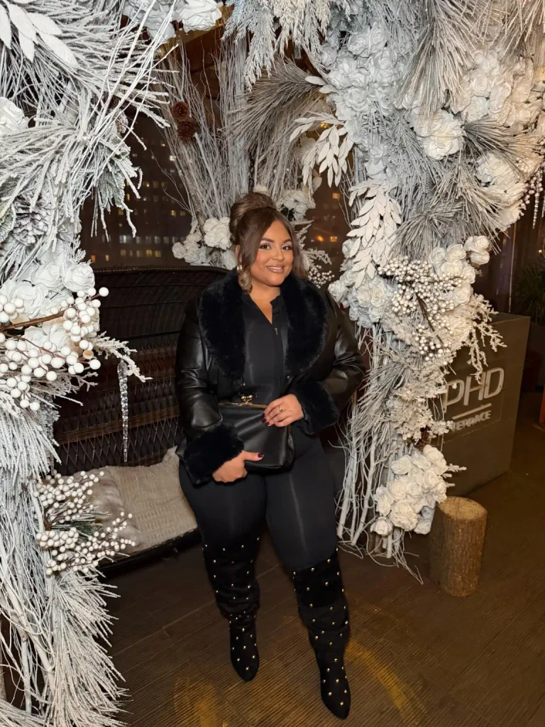 Plus-size woman styling black studded over-the-knee boots with fitted black trousers, a faux-fur-trimmed leather-look coat, and a structured black handbag for a stylish winter evening outfit.