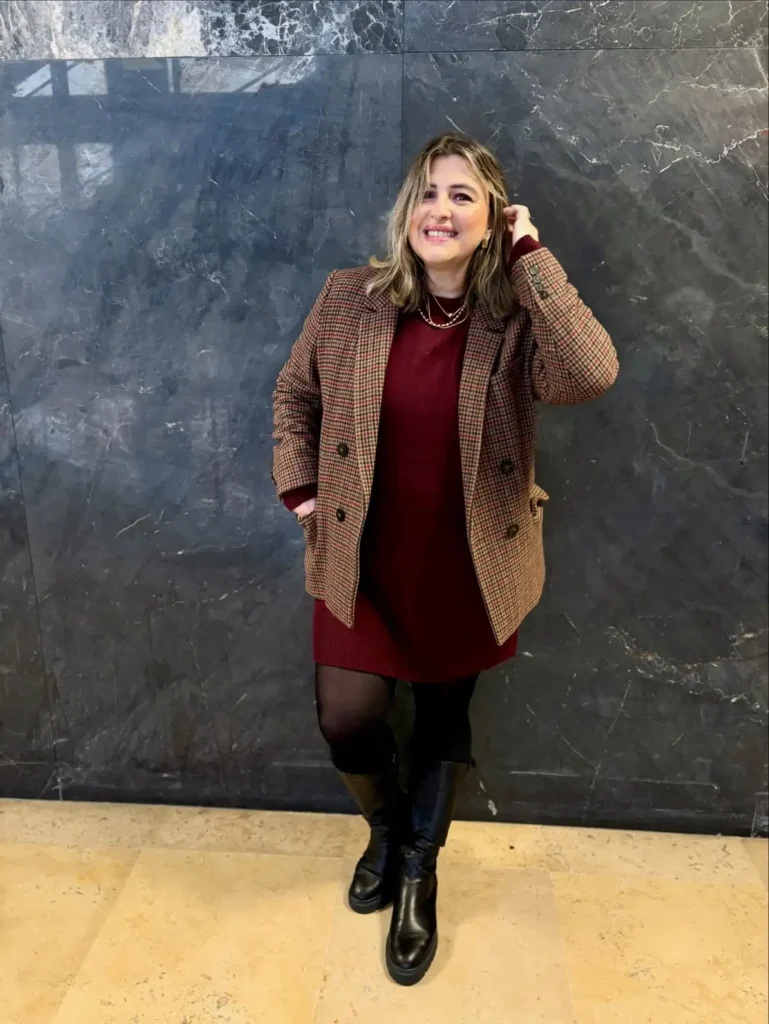  Plus size woman wearing a burgundy knit mini dress with a plaid blazer, black tights, and tall black boots for a classic fall outfit.