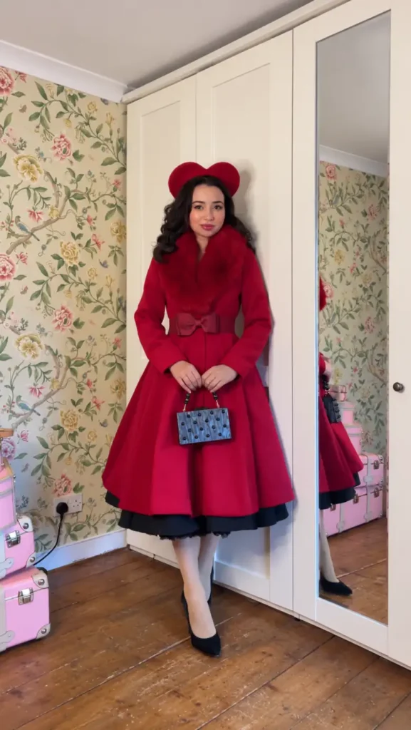 Woman wearing a bright red flared winter coat with a plush collar, bow waist belt, matching red hat, black heels, and a small handbag.