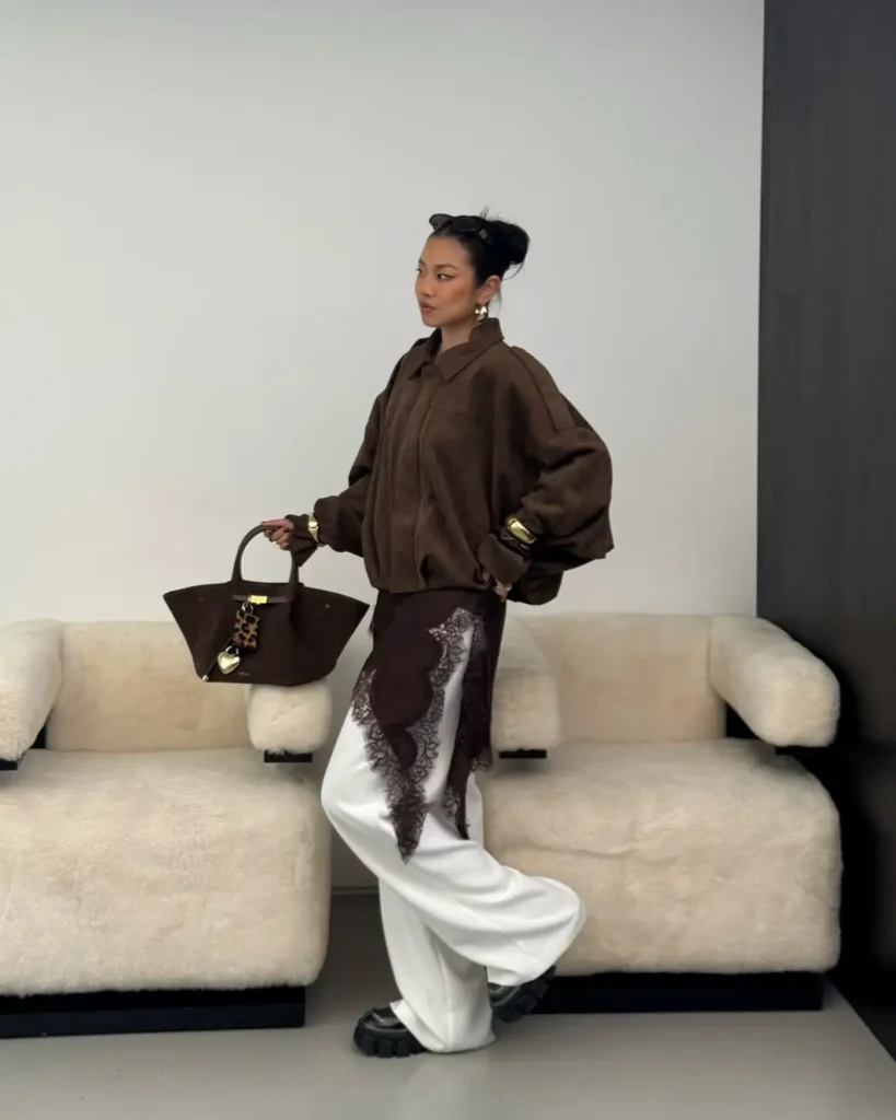 Woman wearing a brown oversized bomber jacket with white wide-leg trousers and an asymmetrical dark lace overlay.