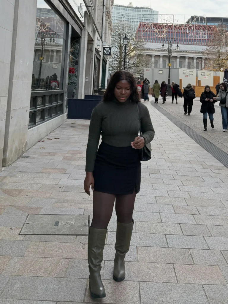 Black woman wearing a fitted olive green ribbed sweater with a black mini skirt, sheer tights, tall olive boots, and a black shoulder bag for a simple fall city outfit.
