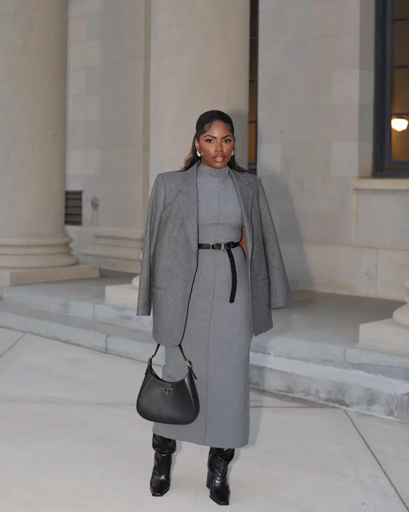Black woman wearing a fitted gray high-neck midi dress with a matching blazer, black waist belt, knee-high boots, and a black shoulder bag for elegant office fashion.