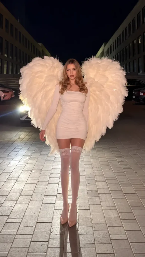 Woman wearing a fitted white mini dress with oversized white feather angel wings, thigh-high stockings, and nude pointed heels.