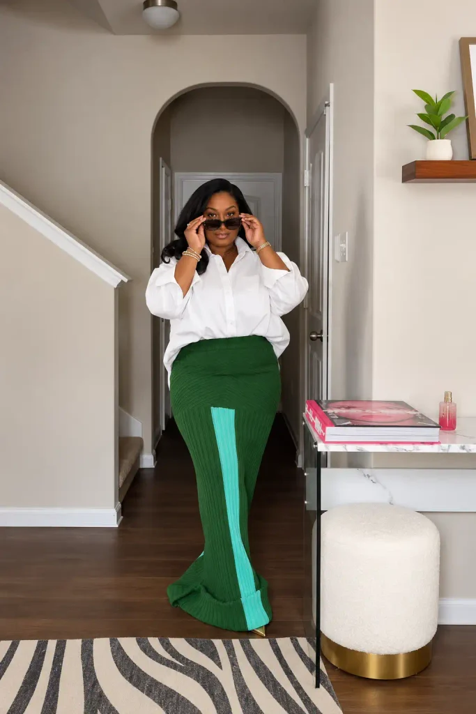 Plus size Black woman wearing an oversized white button-down shirt with an emerald green ribbed maxi skirt featuring a mint-green panel, gold jewelry, and sunglasses for a chic fall outfit.