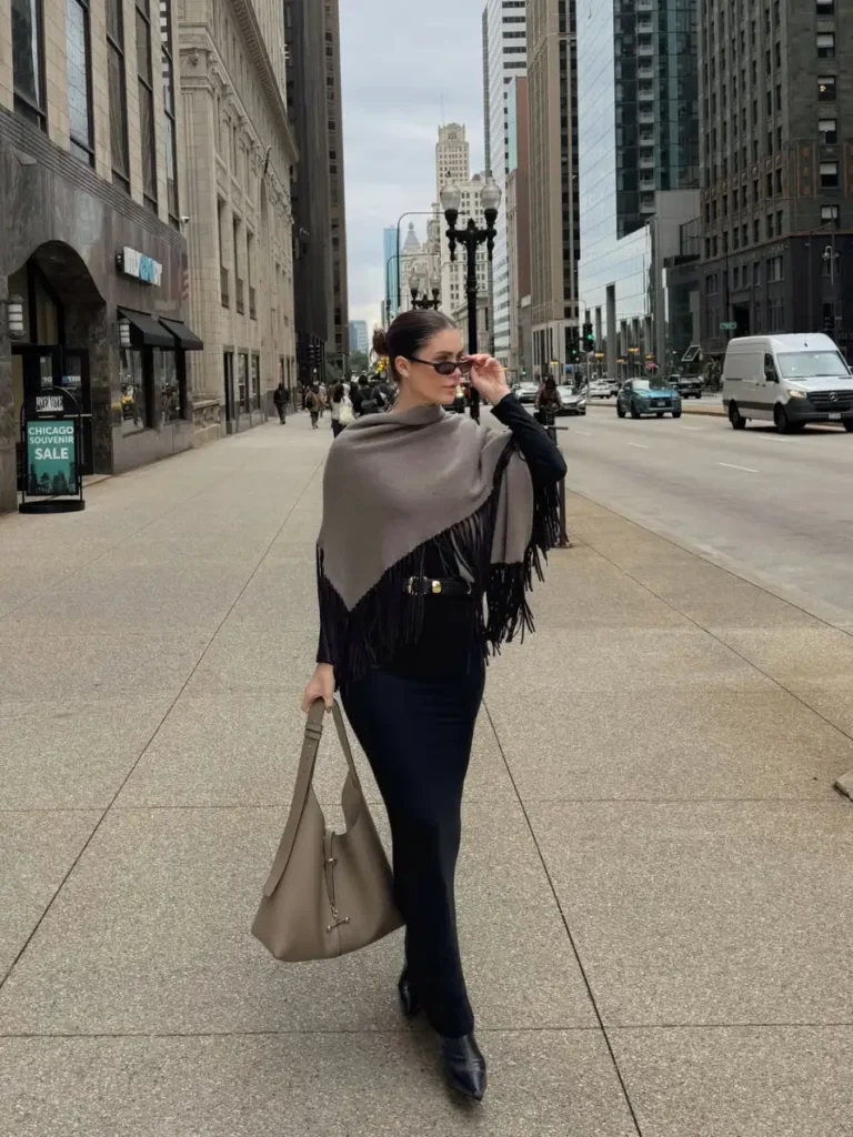 Woman wearing a taupe fringed wrap over a fitted black outfit with a slim belt, pointed black boots, and a large taupe handbag for a sophisticated city fall look.
