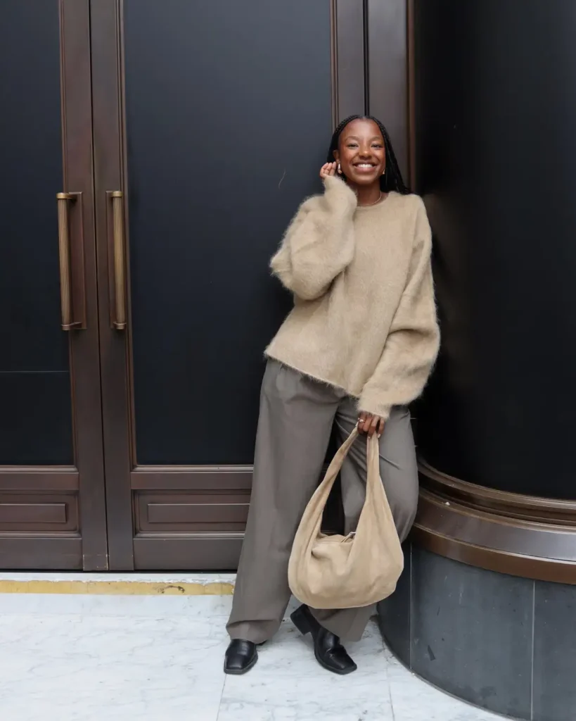 Black woman wearing a fuzzy beige oversized sweater with relaxed gray wide-leg trousers, black square-toe shoes, and a soft beige shoulder bag for a neutral fall outfit.