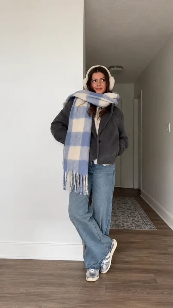 Woman styling a charcoal-gray cropped coat with an oversized blue-and-cream plaid scarf, fluffy cream earmuffs, light-wash wide-leg jeans, and chunky sneakers for a cozy casual winter outfit.