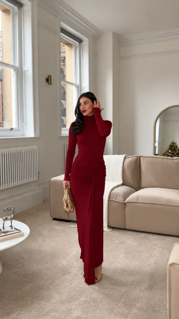 Woman wearing a fitted burgundy long-sleeve maxi dress with a high neckline, gathered waist, draped skirt, metallic gold clutch, and gold heels.