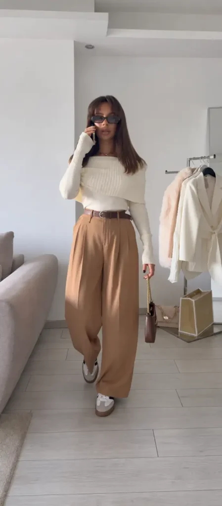 Woman wearing a cream foldover knit sweater with camel pleated wide-leg trousers, a brown belt, retro sneakers, and a small brown bag for a chic casual fall look.