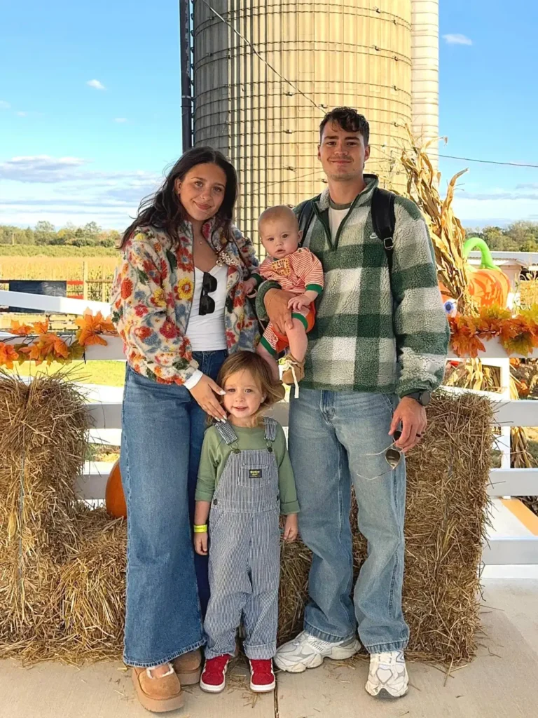 Family wearing casual fall farm outfits with Mom in a colorful floral fleece and denim skirt, Dad in a green checked fleece, a child in striped overalls, and baby in an orange striped outfit.