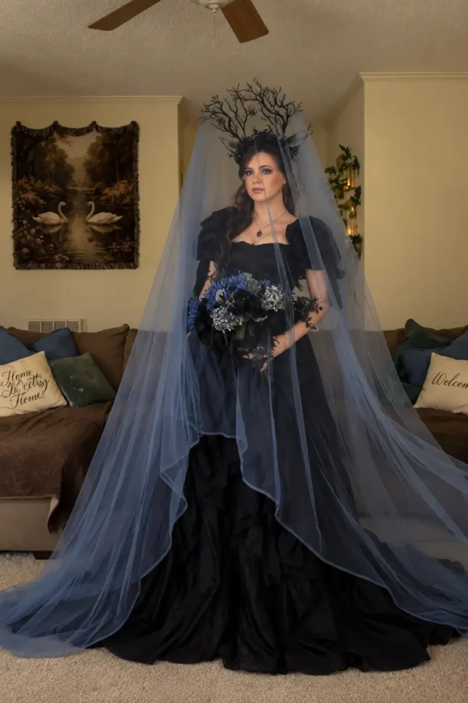Woman wearing a dramatic black gothic bridal gown with a long blue-gray veil, branch crown, and dark blue and black flower bouquet.
