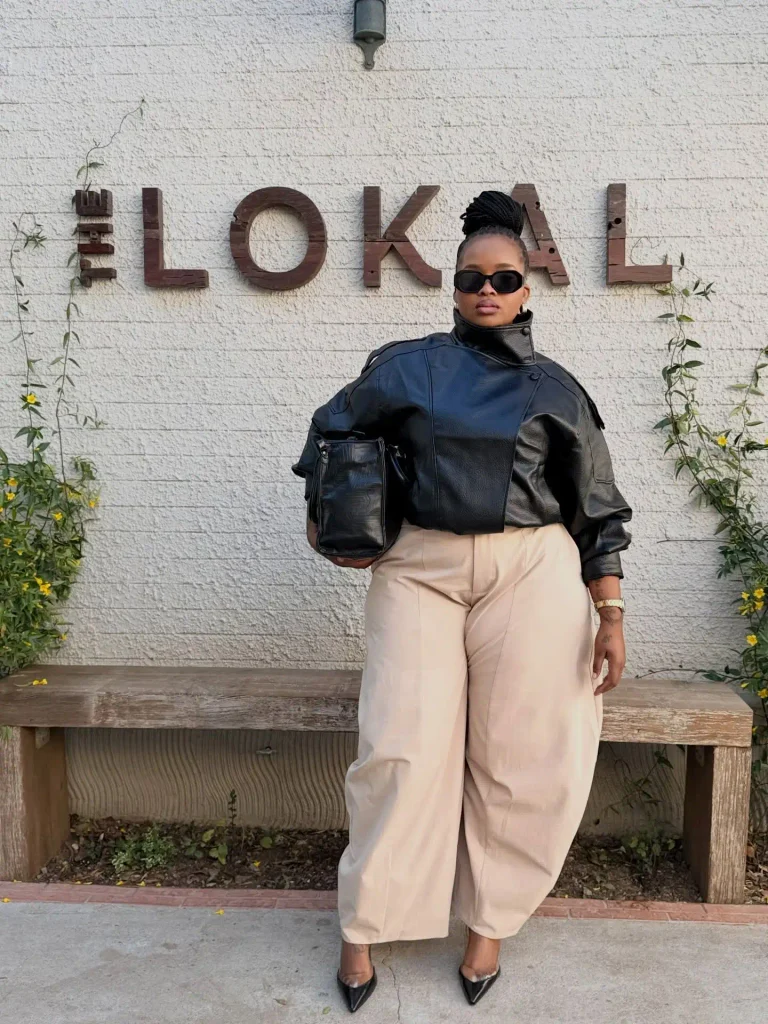 Curvy Black woman wearing an oversized black leather bomber jacket with beige barrel-leg trousers, pointed black heels, sunglasses, and a structured handbag for trendy fall street style.