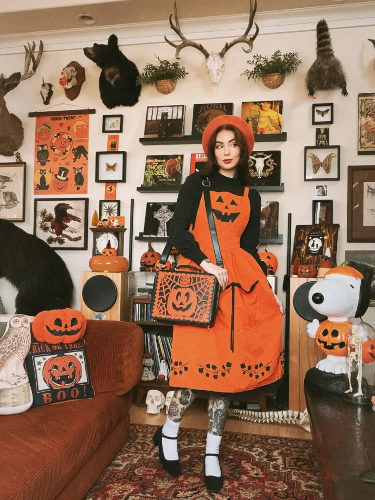 Woman wearing an orange jack-o-lantern pinafore dress over a black blouse with orange beret, white socks, black shoes, and pumpkin bag.