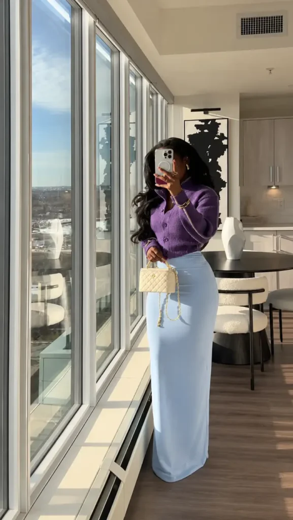 Black woman wearing a cropped purple button-up cardigan with a light blue maxi skirt, a cream quilted handbag, and gold jewelry for a feminine, colorful winter outfit.