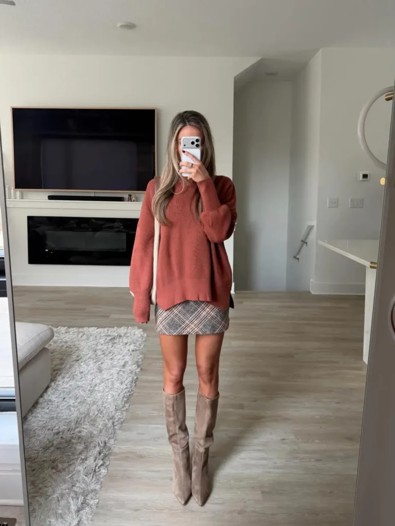 Woman wearing an oversized rust-colored sweater with a brown and gray plaid mini skirt, taupe suede knee-high boots, and a cream shoulder bag for a cozy Thanksgiving outfit.