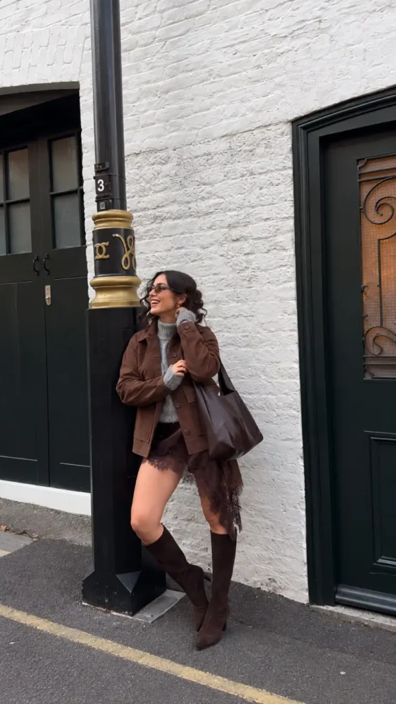 Woman wearing a brown jacket with a grey knit sweater, asymmetrical brown lace skirt detail, knee-high boots, and a large brown bag.