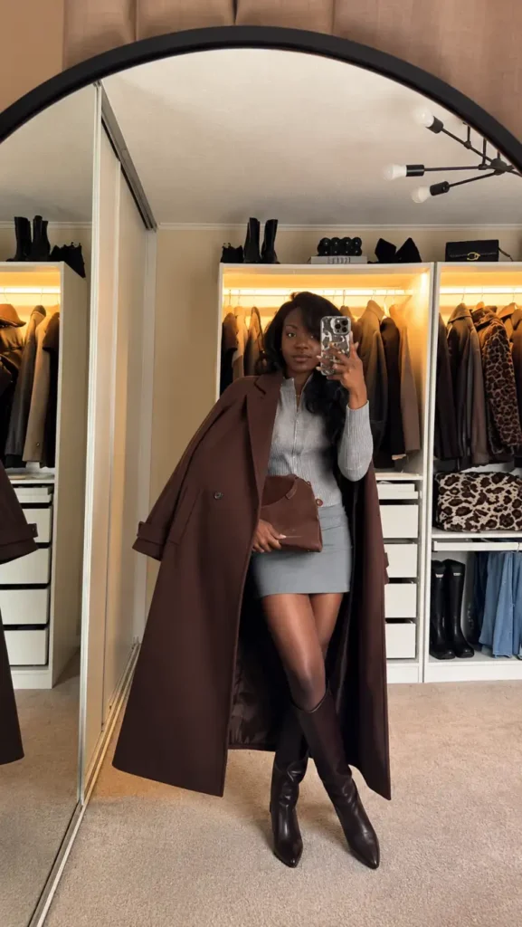 Black woman wearing a gray fitted mini dress with a long chocolate brown coat, knee-high brown boots, and a matching clutch for a chic fall outfit.