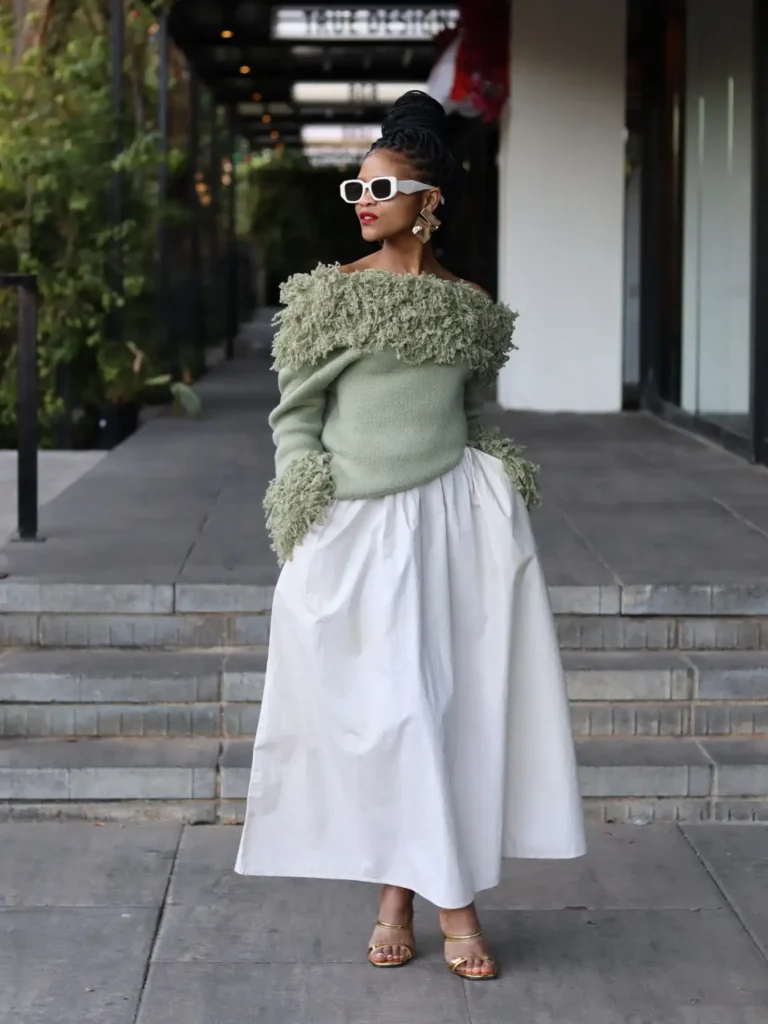 Stylish Black woman wearing a sage green off-shoulder fringe sweater with a voluminous white maxi skirt, white sunglasses, gold earrings, and gold sandals.