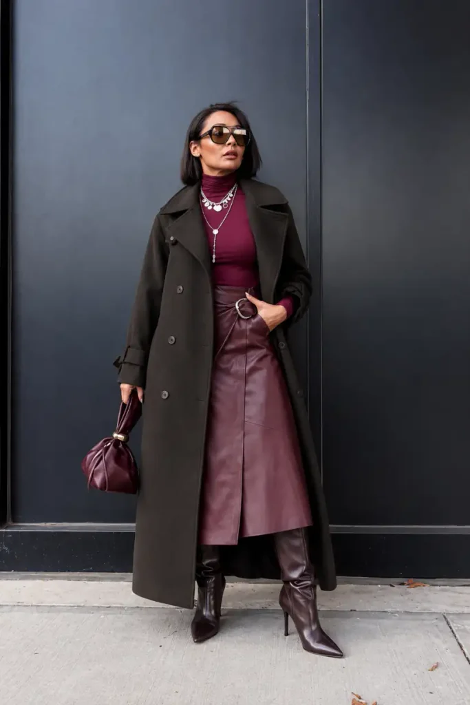 Woman wearing a burgundy turtleneck, leather-look midi skirt, long dark coat, and knee-high boots with a matching handbag for a classy autumn look.