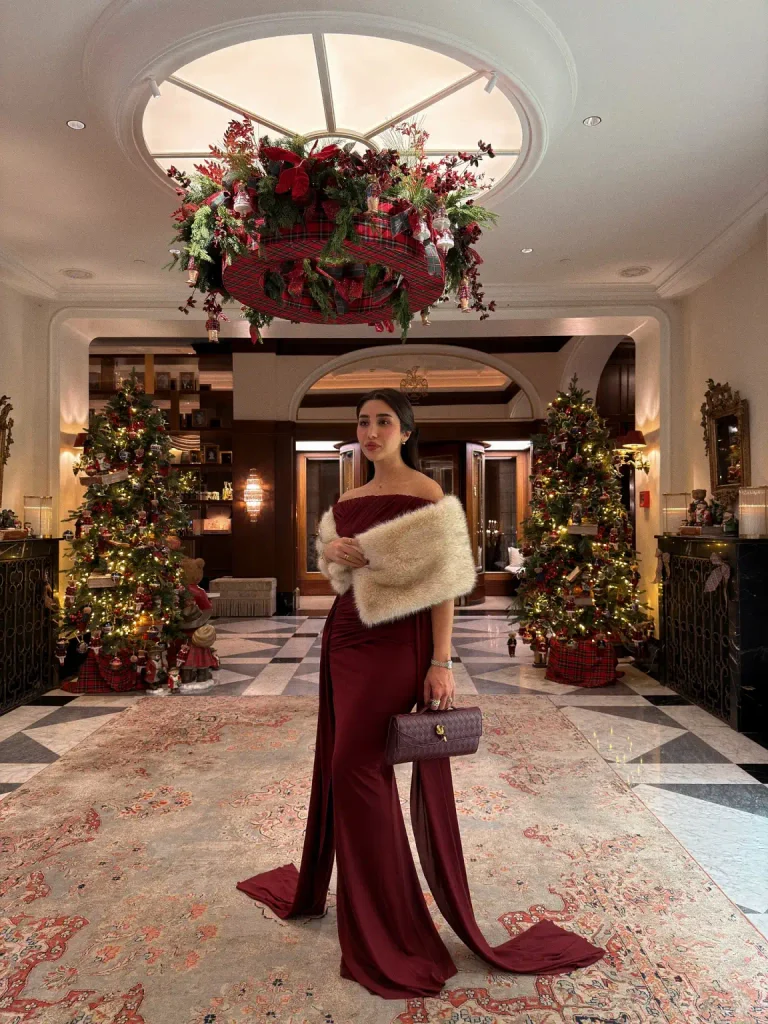 Woman wearing a burgundy off-shoulder evening gown with elegant draping, a cream faux-fur wrap, and a matching quilted clutch for a formal Christmas Eve look.