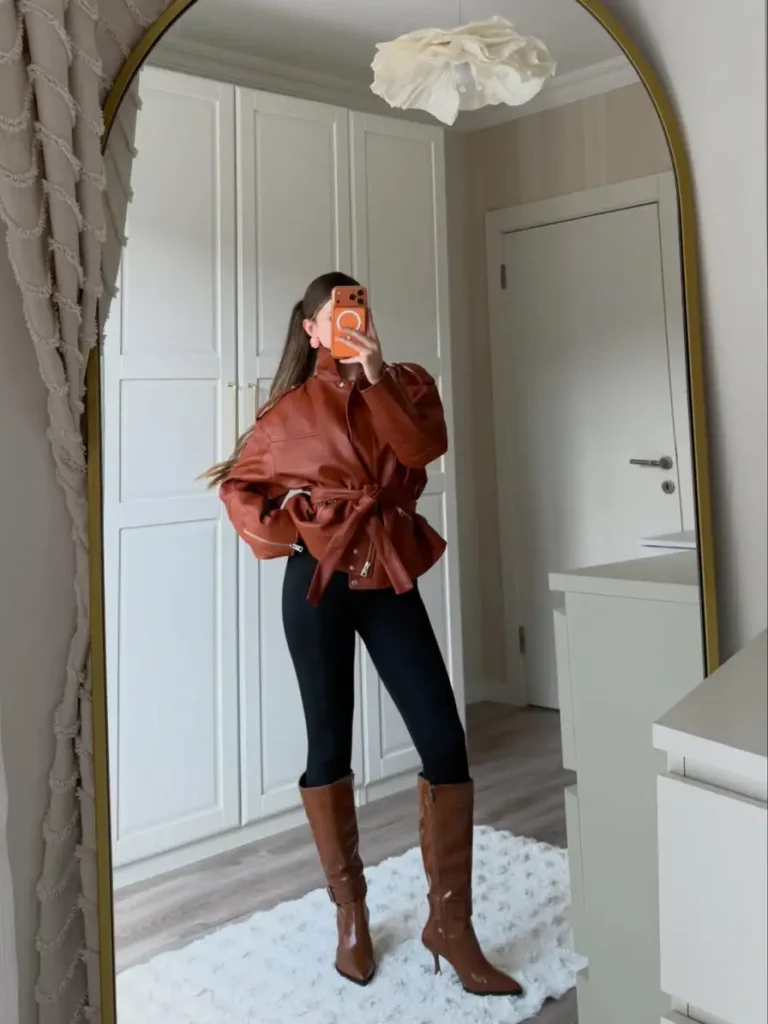 Woman wearing an oversized cinnamon brown leather jacket with black leggings and tall brown boots for a sleek casual fall outfit.