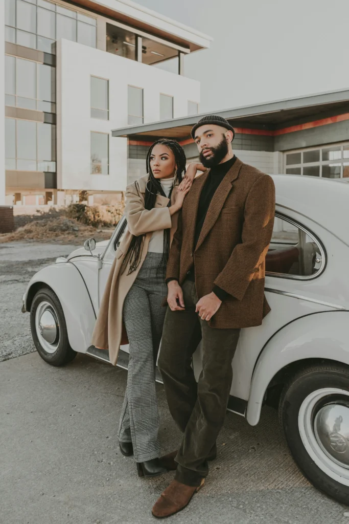 Couple wearing coordinating fall outfits with a camel coat, cream turtleneck, plaid trousers, brown checked blazer, and black knit top for Thanksgiving.