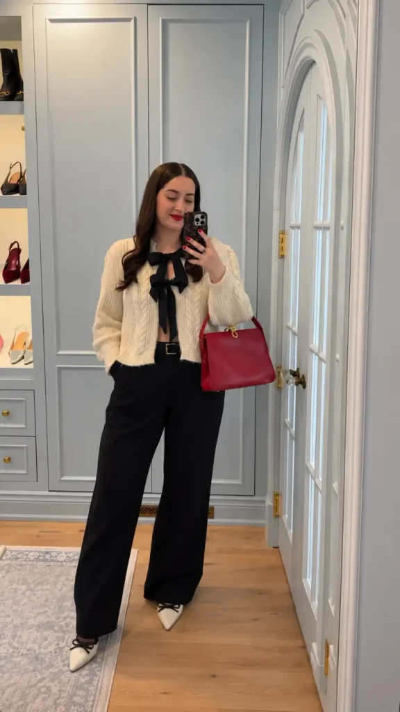 Woman styling a cream cable-knit bow cardigan with black wide-leg trousers, white pointed heels, and a red handbag for a classy holiday outfit.
