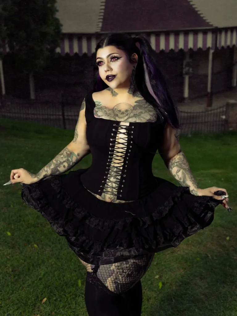 Plus size woman in a black lace-up corset, ruffled skirt, fishnet tights, and dark vampire makeup for Halloween.