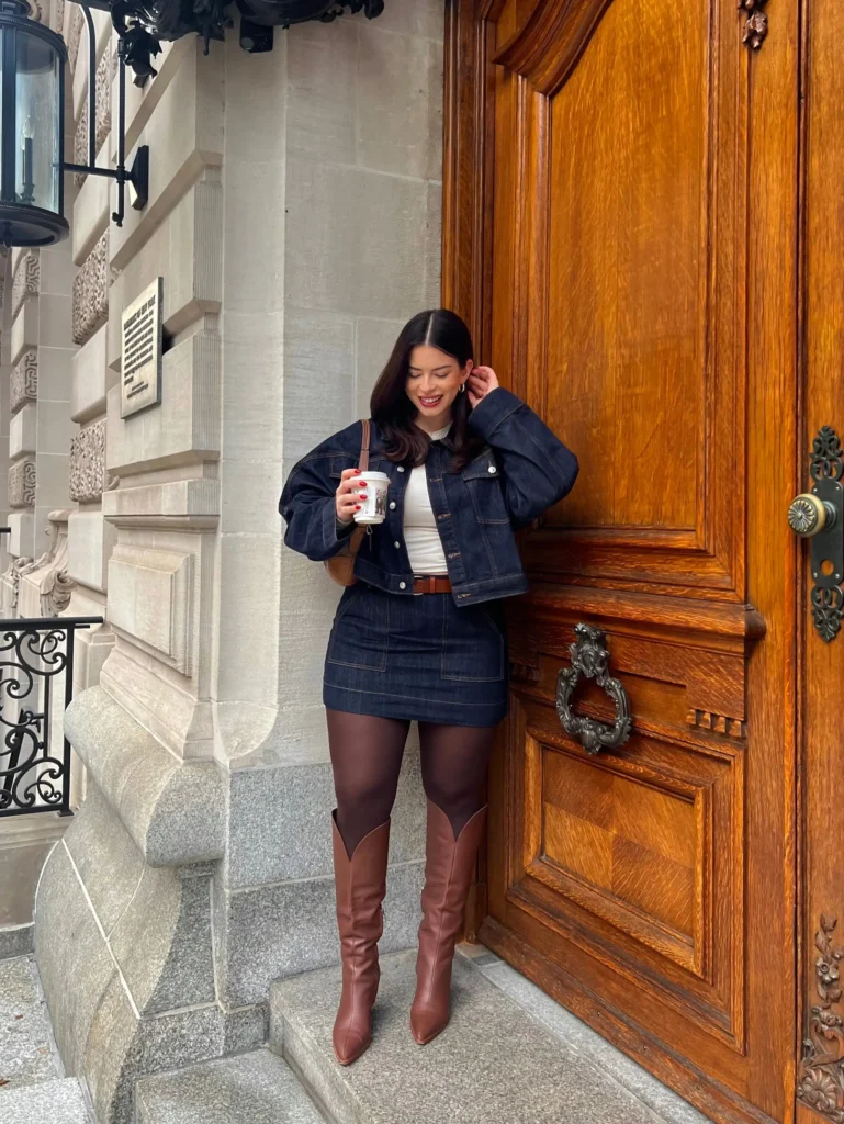 Woman wearing dark denim jacket and matching mini skirt with sheer tights and brown knee-high boots.