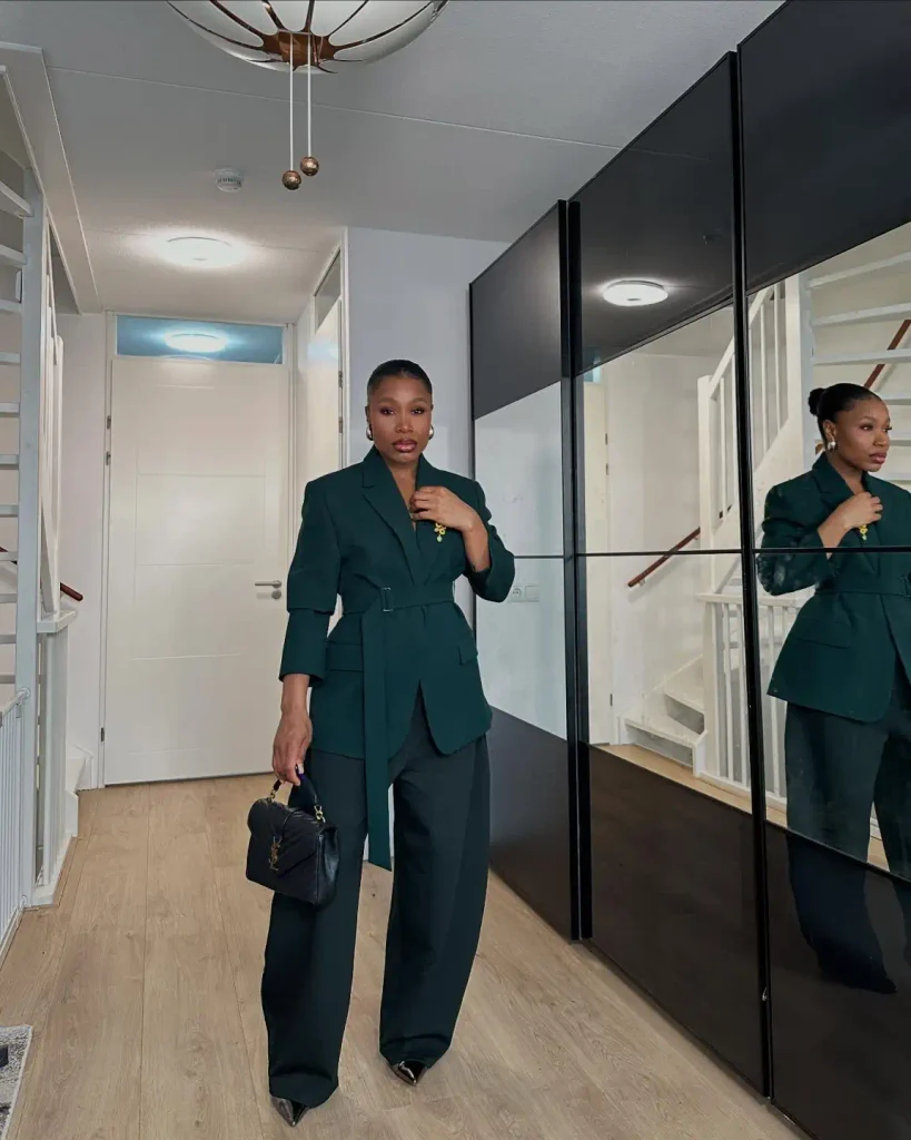 Black woman wearing a forest green belted blazer with matching wide-leg trousers, pointed heels, gold earrings, and a black handbag for a powerful corporate outfit.