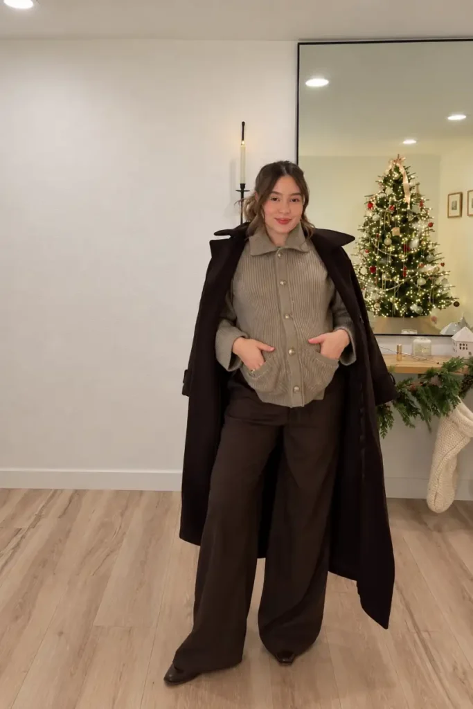 Pregnant woman wearing a taupe ribbed cardigan, chocolate brown wide-leg trousers, and a long dark coat for a stylish fall maternity outfit.