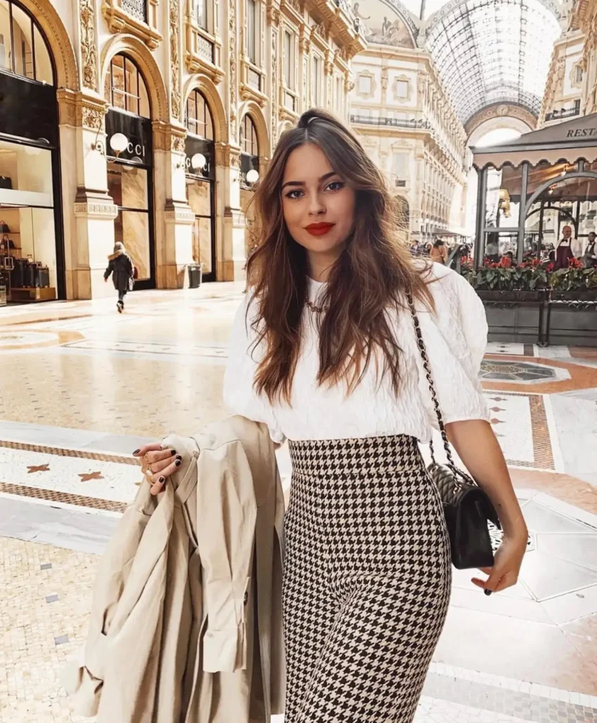Petite woman wearing a white puff-sleeve top with high-waisted houndstooth trousers, a black shoulder bag, and a neutral trench for a classy fall look.
