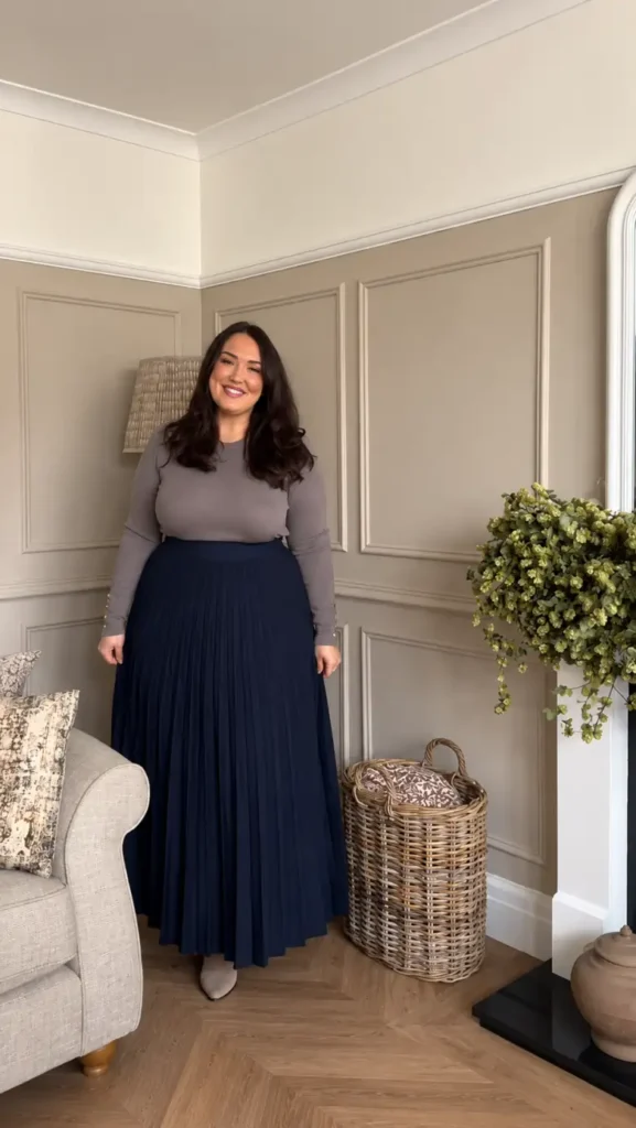 Plus size woman wearing a taupe long-sleeve top with a navy pleated maxi skirt and neutral ankle boots for an elegant fall look.