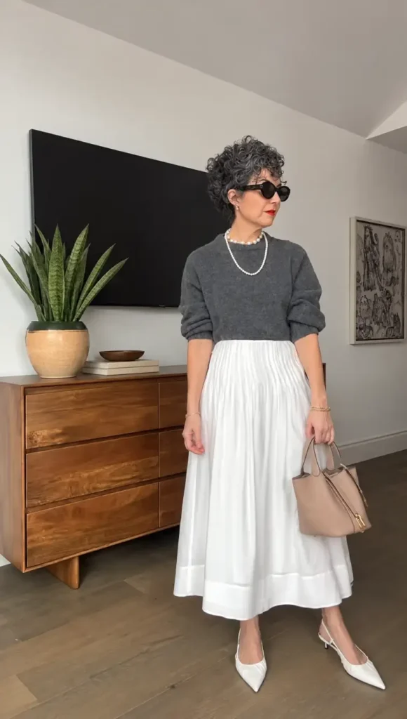 Mature woman wearing a charcoal gray sweater with a flowing white pleated maxi skirt, pearl necklace, and white pointed heels for a classy Thanksgiving dinner outfit.