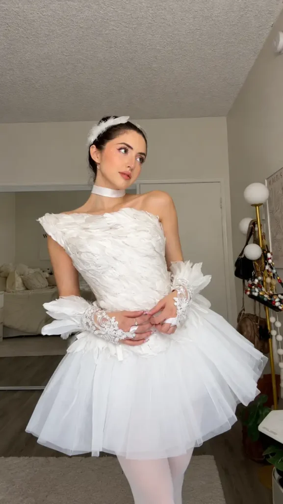 Woman wearing a white feathered ballerina dress with layered tutu, white tights, lace gloves, and feather hairpiece for an elegant Halloween costume.