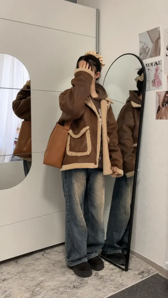 School girl wearing an oversized chocolate-brown shearling jacket over a brown hoodie, baggy faded jeans, chunky dark shoes, and a tan shoulder bag for a casual winter outfit.