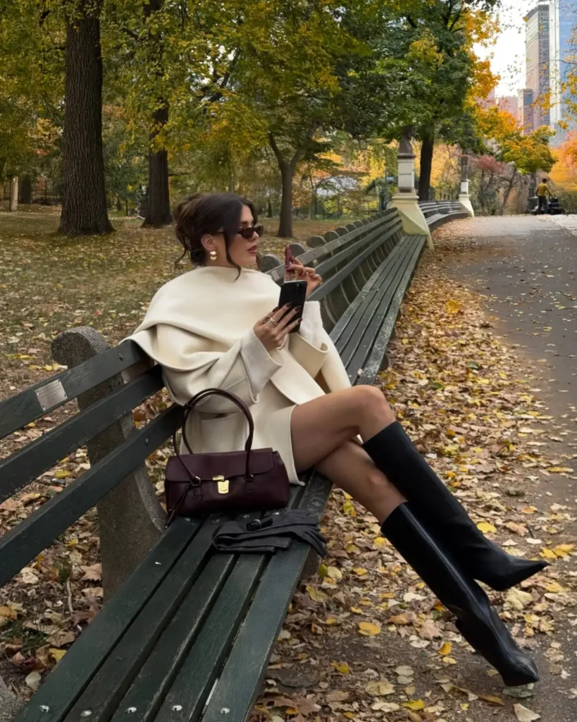 Cream cape styled with a neutral mini outfit, black knee-high boots, sunglasses, and a burgundy handbag for an elegant autumn outfit.