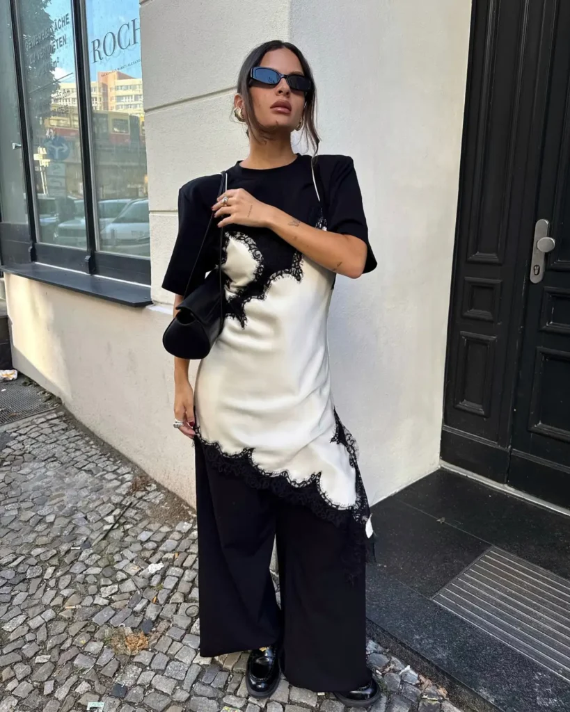 Woman layering an ivory lace-trim slip dress over black wide-leg trousers with a black top, chunky shoes, and shoulder bag.