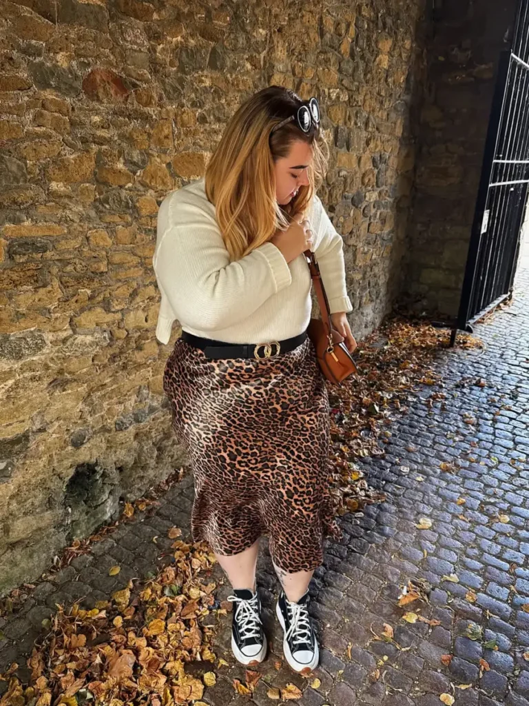 Plus size fall outfit featuring a white knit sweater tucked into a leopard print midi skirt with a black belt, high-top sneakers, and brown crossbody bag.