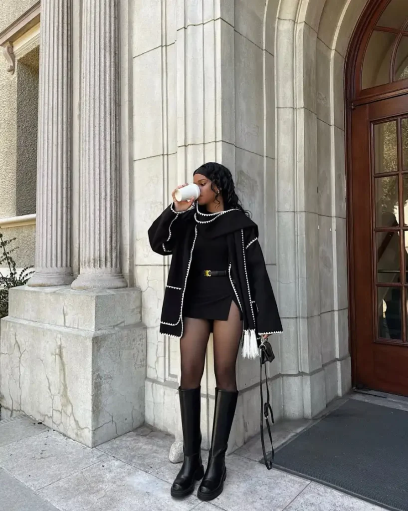 Black woman wearing a black mini skirt and fitted black top with an oversized black jacket trimmed in white, tall black boots, and a small black bag for a polished fall outfit.