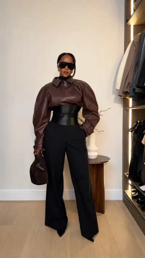 Black woman wearing a chocolate brown leather-look top with a wide black corset belt, black wide-leg trousers, pointed heels, and a dark brown handbag for stylish fall workwear.