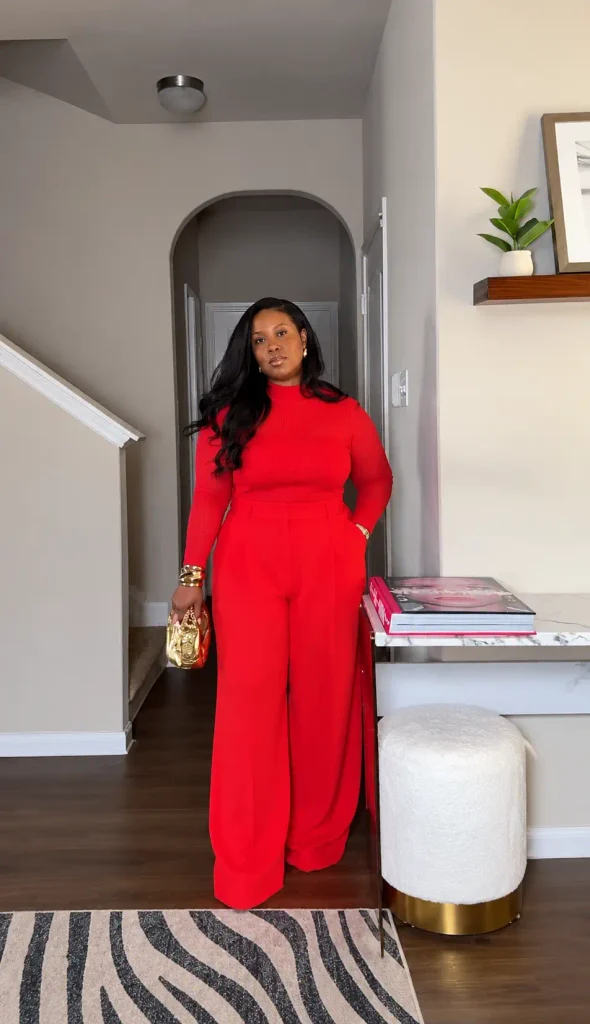 Plus size Black woman wearing a bright red ribbed turtleneck with matching high-waisted wide-leg trousers, a metallic gold clutch, and stacked gold bracelets for a bold fall outfit.