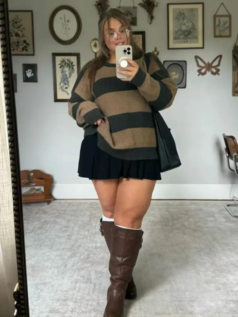 Plus size woman wearing an oversized brown and black striped sweater with a black pleated mini skirt, tall brown boots, and a black tote for fall.