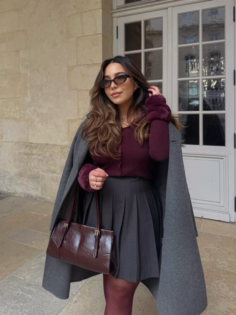 Burgundy cardigan styled with a charcoal pleated mini skirt, long gray coat, burgundy tights, and matching handbag for a chic fall outfit.